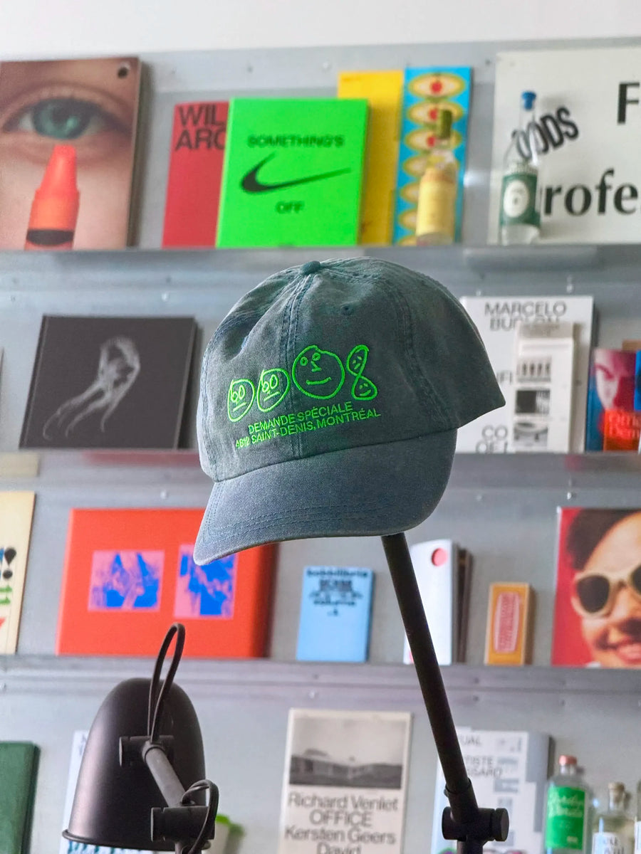 Casquette bleu-verte Demande Spéciale sur un présentoir, devant une étagère de livres de design