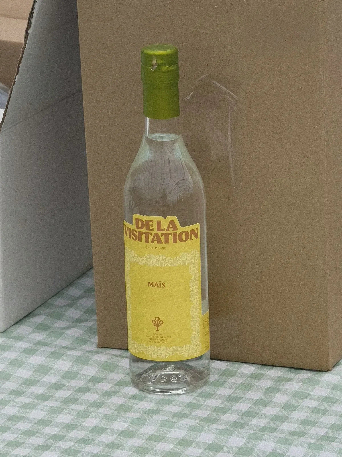 De la Visitation — bouteille à étiquette jaune posée devant une boîte, nappe à carreaux verts