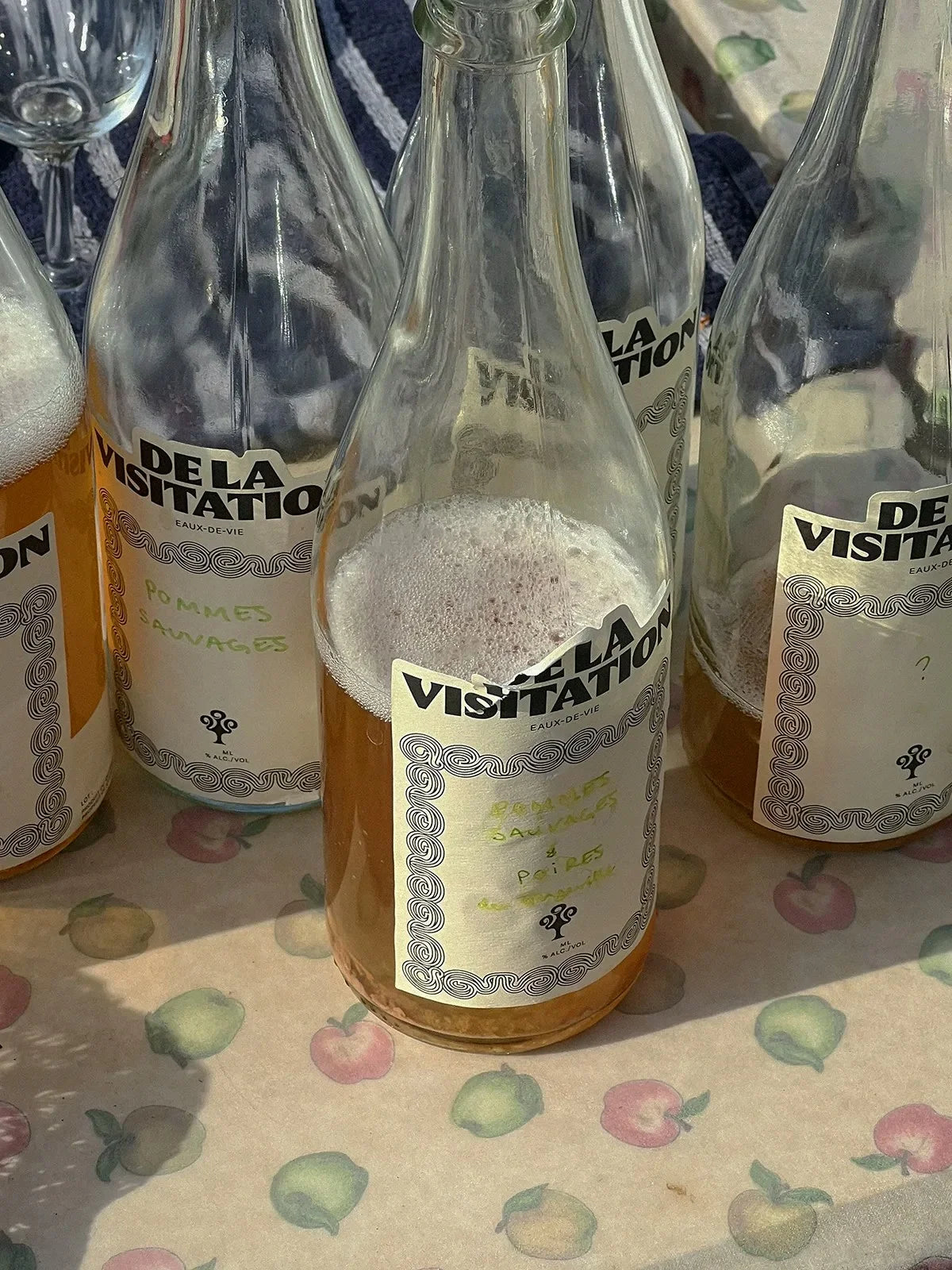 De la Visitation — bouteilles d'eau-de-vie aux étiquettes blanches alignées sur une surface fleurie