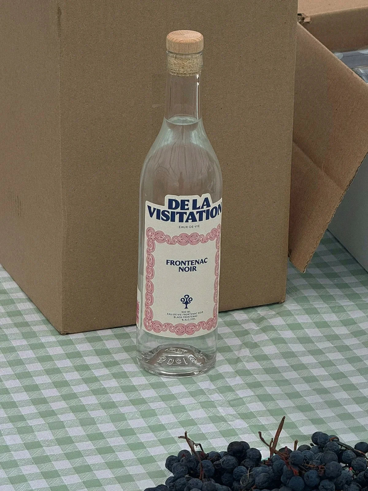 De la Visitation — bouteille à liseré rose devant une boîte, raisins noirs et nappe à carreaux