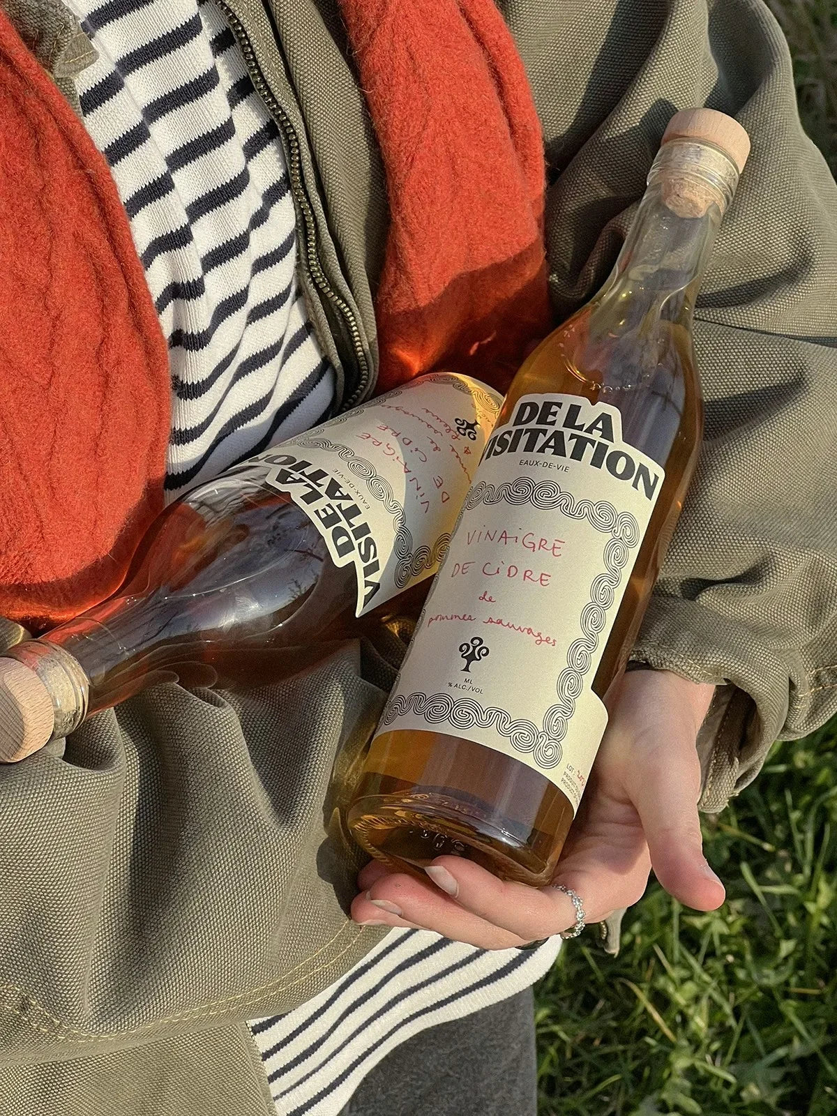 De la Visitation — main tenant deux bouteilles ambrées en extérieur, manteau kaki et rayé