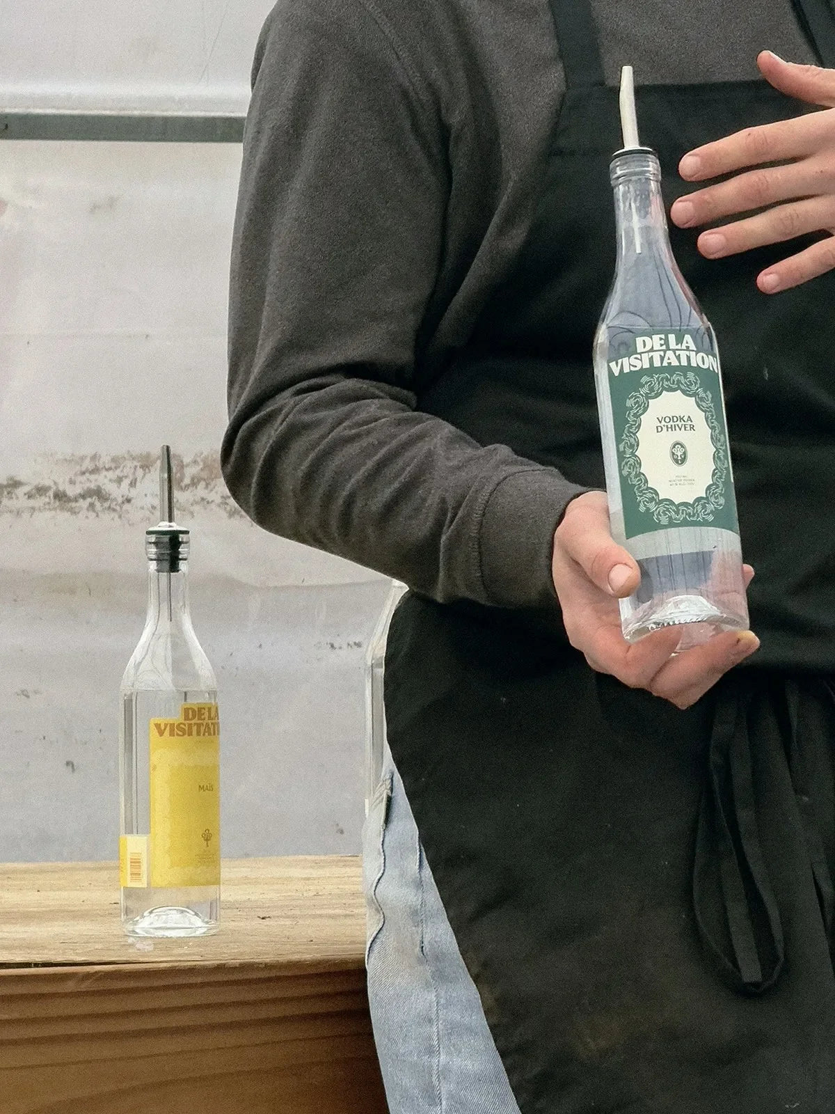 De la Visitation — personne en tablier tenant une bouteille à étiquette verte près d'un comptoir