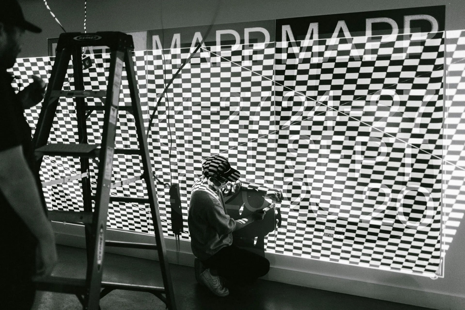  L'inconnu — technicien agenouillé devant une projection murale au motif optique noir et blanc, montage en cours