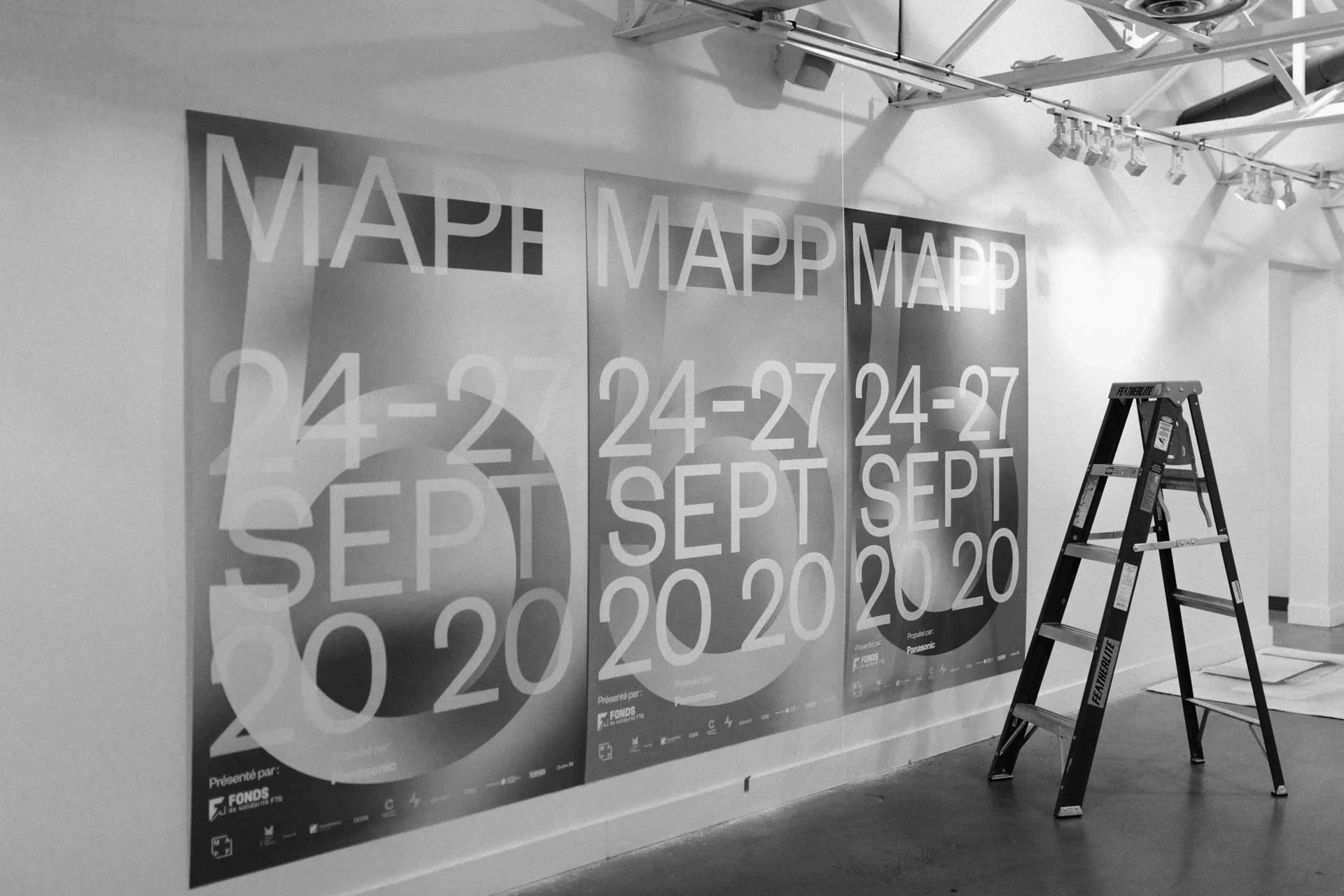 L'inconnu — trois affiches MAPP géantes au mur d'une galerie en montage, escabeau à droite, photo noir et blanc