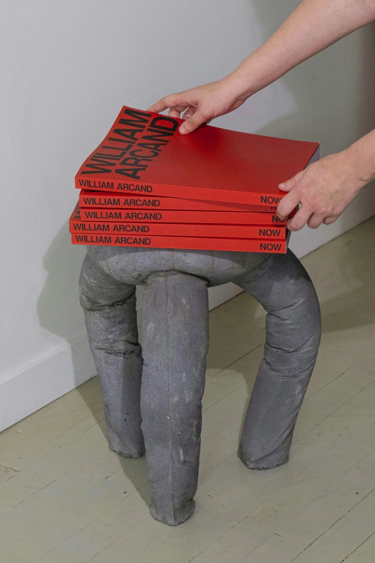 Now — mains posant une pile de livres rouges « William Arcand » sur un tabouret de pierre sculpté