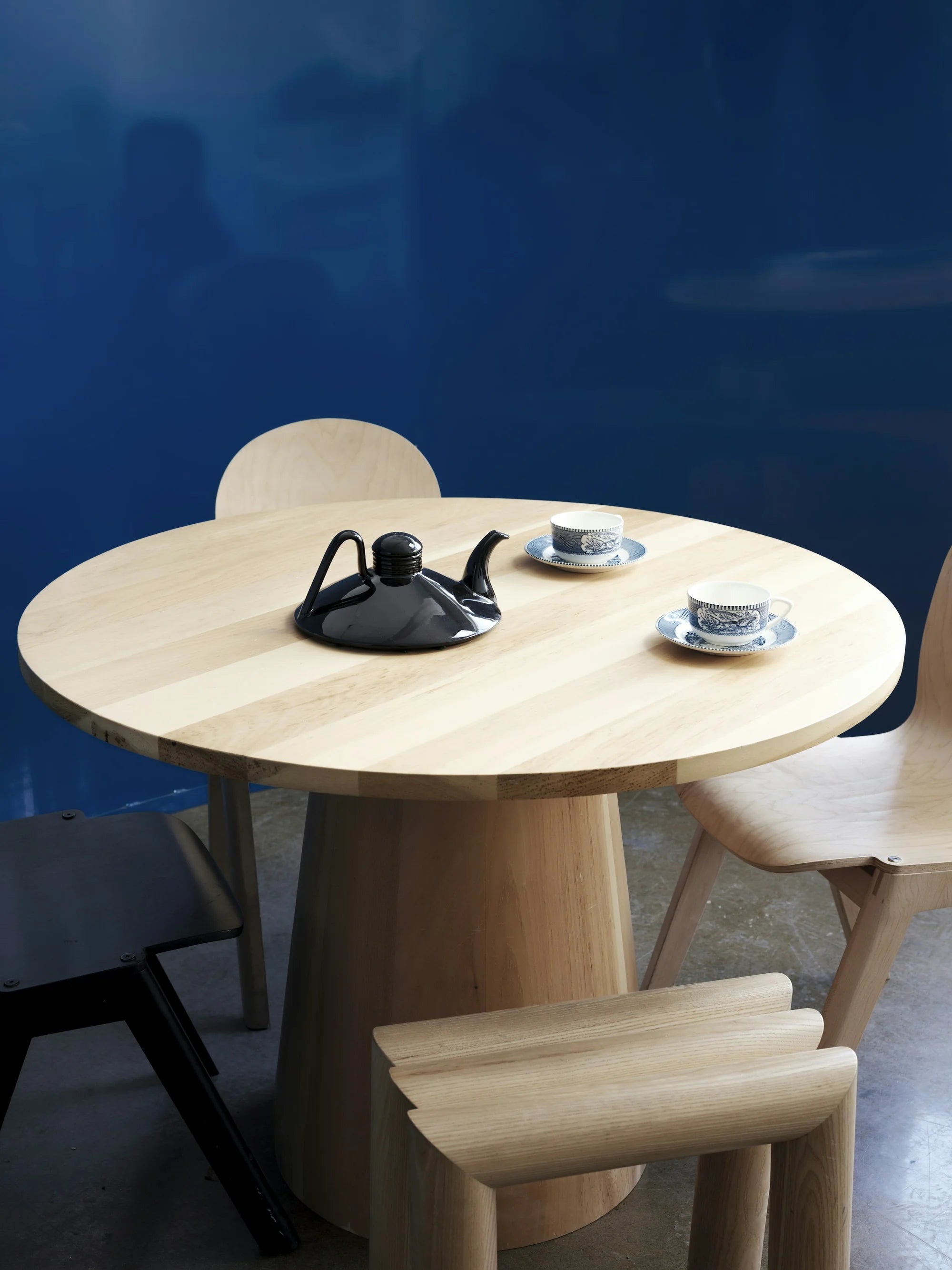 SANGARE — table ronde en bois clair avec théière noire et tasses, mur bleu profond en arrière-plan