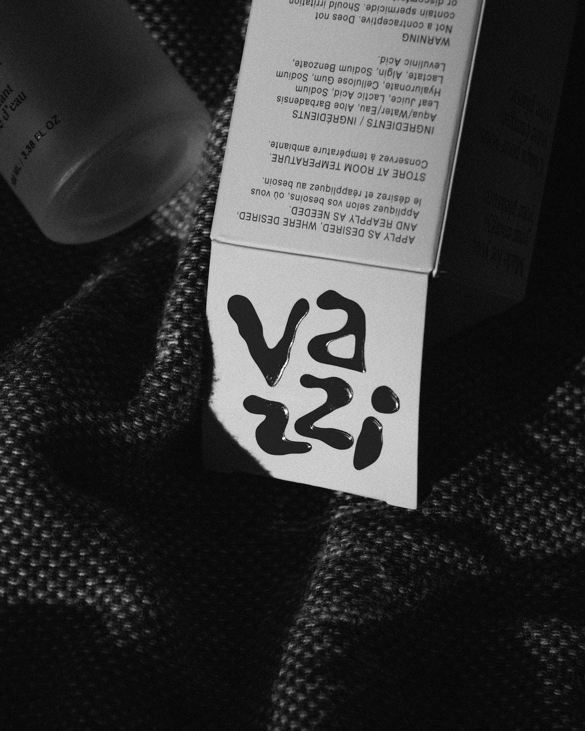 Vazzi — étiquette de tissu au logo bleu manuscrit, flacon en arrière-plan, noir et blanc