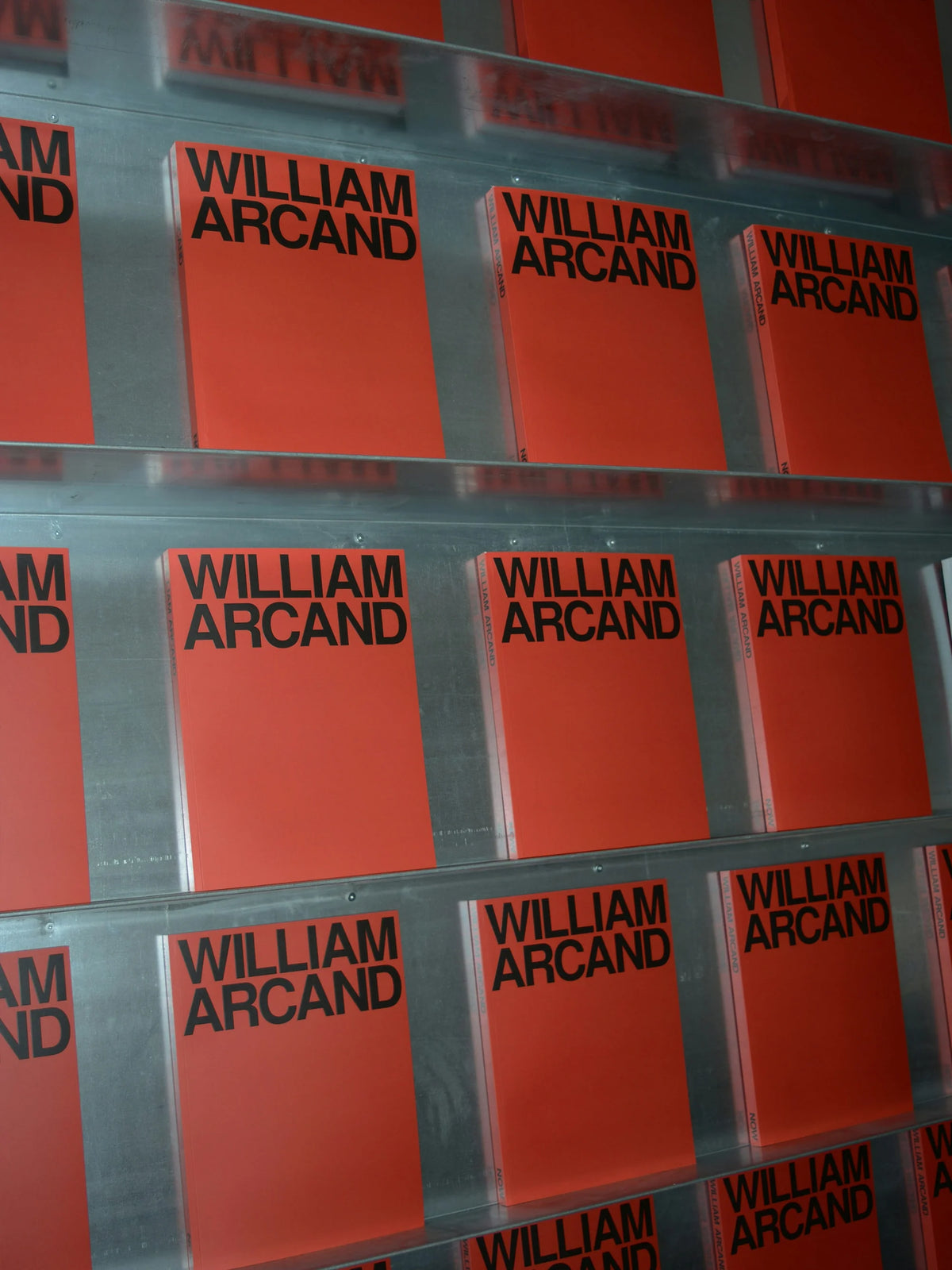 Now — couvertures rouges du livre « William Arcand » alignées en gros plan sur des tablettes