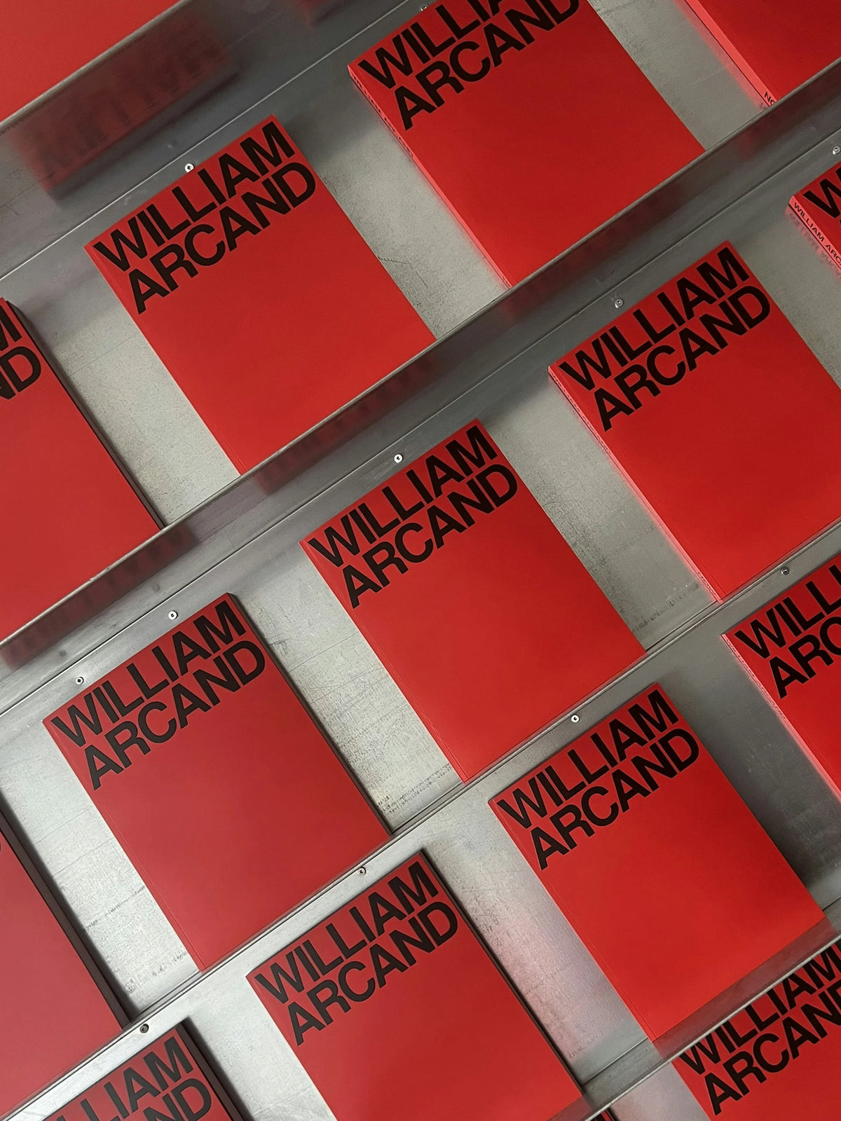 Now — grille d'exemplaires à couverture rouge au wordmark noir « William Arcand » sur étagères